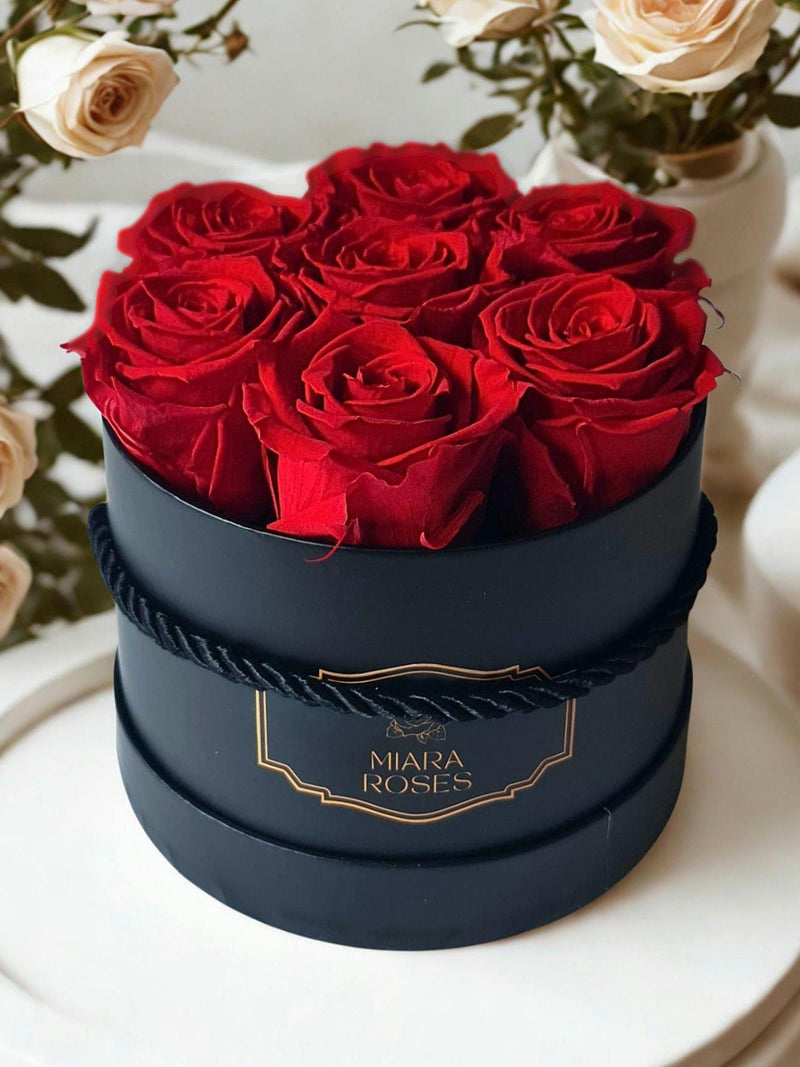 Dark gift box with 7 preserved red roses and gold logo label, tied ribbon