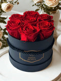Dark gift box with 7 preserved red roses and gold logo label, tied ribbon