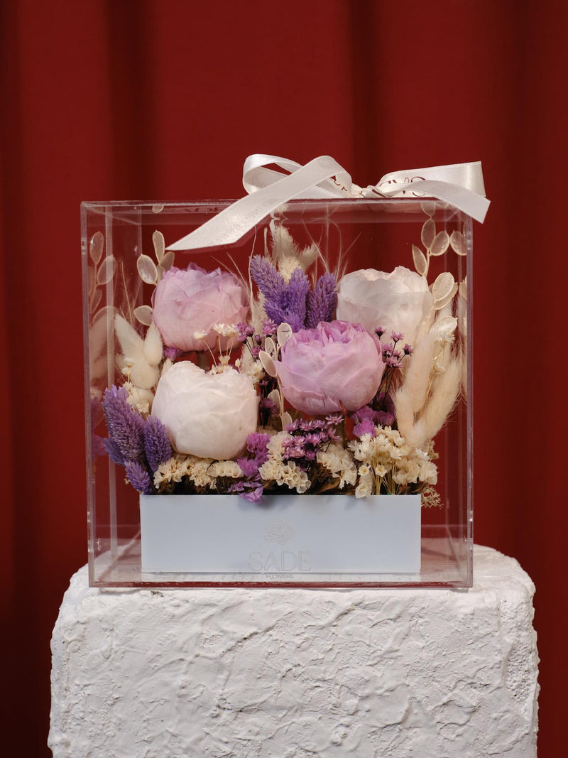 Preserved peony floral centerpiece in clear acrylic box with ribbon, purple and pink blooms