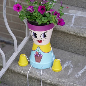 Decorative girl flower pot with face graphic, blue base and purple rim, holding purple flowers