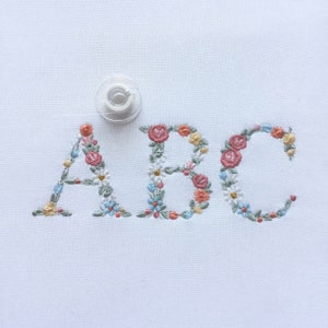 Floral embroidered uppercase alphabet letters, blue and pink flower accents on white fabric
