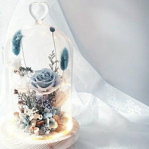 Blue preserved rose and dried flowers under clear glass dome with warm base light