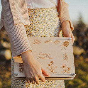 Personalized flower press with botanical line-art cover, held in hands outdoors