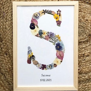 Framed pressed-flower initial with colorful dried blooms on white background, with small text