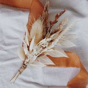 Dried flower and feather boutonniere with warm orange and cream accents, pinned lapel.