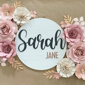 Round wood name sign reading Sarah JANE with pink mauve paper flowers and leaf accents