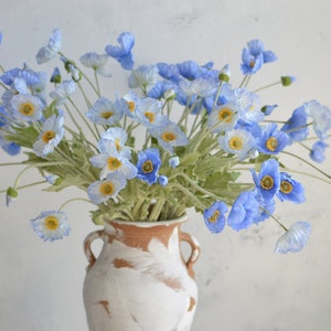 Faux blue poppy-style flower heads with white daisies and green stems, in vase