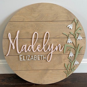 Round wooden nursery name sign with floral snowdrop design and text 'Madelyn Elizabeth'