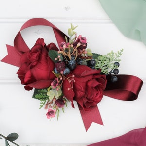 Artificial burgundy rose wrist corsage with navy ribbon, greenery, and small blooms.