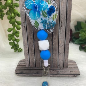 Blue pressed-flower badge reel with clear resin oval, beads, and metal clip