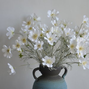 Artificial white cosmos flower branch with small daisy blooms and yellow centers in a dark vase