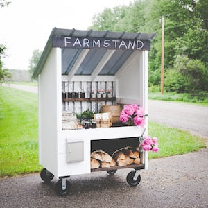 White farmstand cart with pitched roof, open shelves, and wheel base