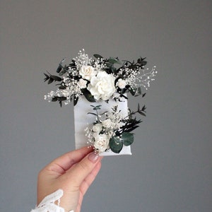 Boutonniere with white flowers, green leaves, and small dark accents on a pinned backing