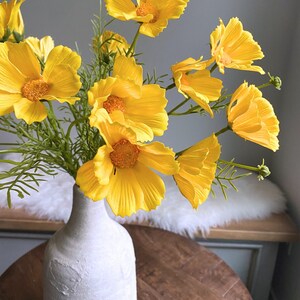 Yellow artificial wildflower stem with daisy-like blooms and green leaves in white vase