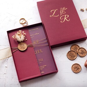 Burgundy wedding invitation sleeve with gold foil monogram, wax seal and dried flowers