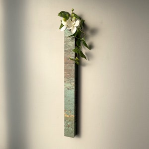 Wall-mounted long vase with single stem of white flowers and green leaves