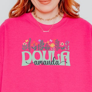 Pink crewneck sweatshirt with colorful floral “Doula” graphic and ribbon text