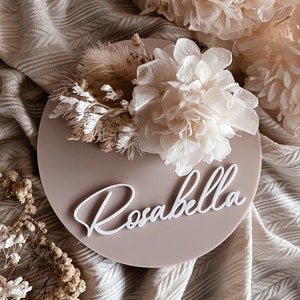 Round acrylic birth announcement sign with cursive “Rosabella” and dried white flowers