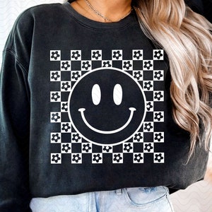 Dark crewneck shirt with smiling face graphic over black-and-white checkered pattern
