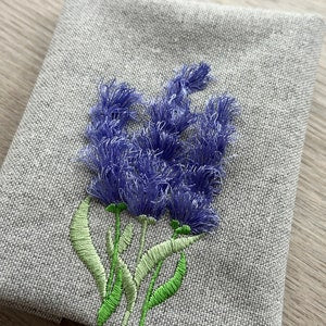 Embroidered lavender-blue flower stem with fluffy petals and green leaves on light fabric