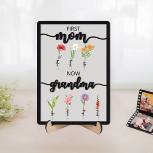 Standing wooden sign with “First mom now grandma” text and colorful birth flowers