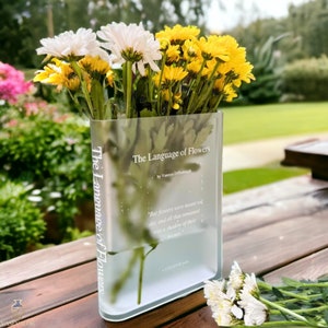 Clear acrylic book-shaped vase holding white and yellow flowers, visible front text