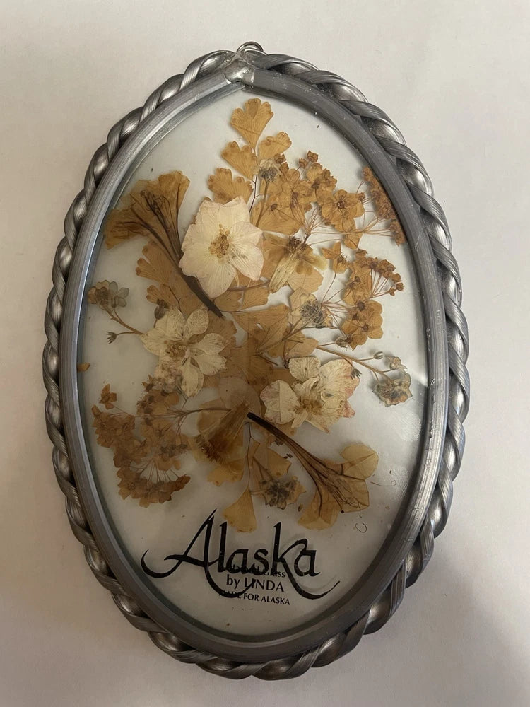 Oval glass frame with pressed dried flowers and 'Alaska' text, metal border
