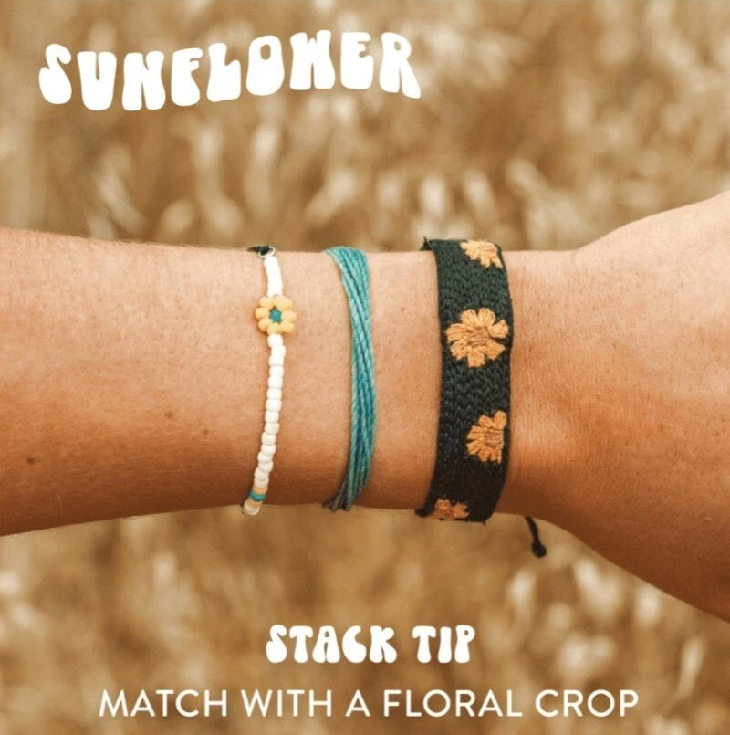 Stacked sunflower bracelets: white bead chain, black woven band with orange sunflowers, teal cord