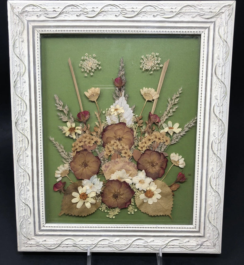 Distressed white framed pressed flowers artwork with dried blooms and greenery on green background