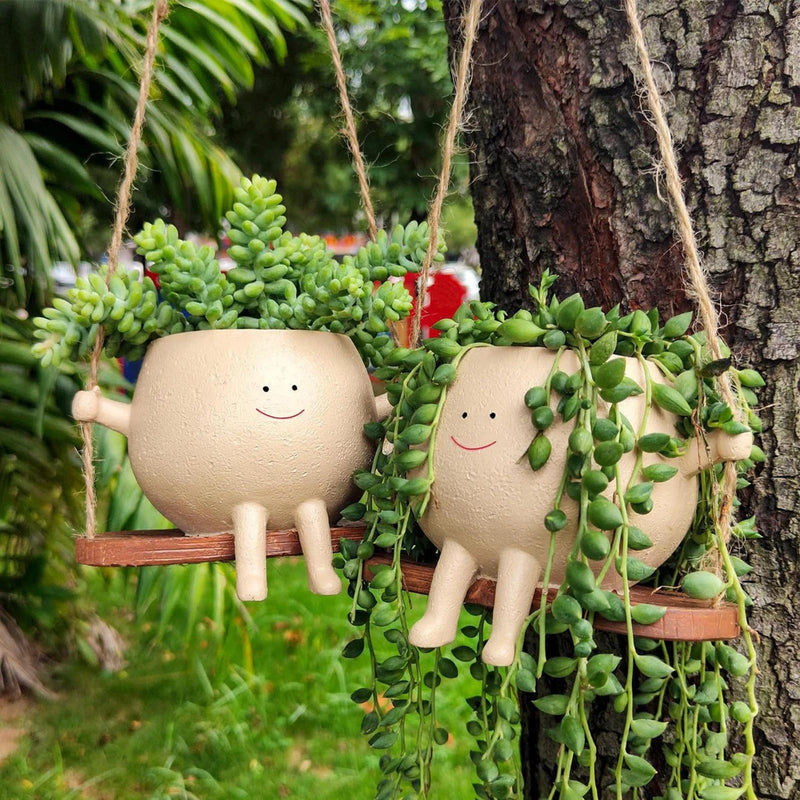 Hanging swing planter pots with smiling faces, holds trailing green succulent plants