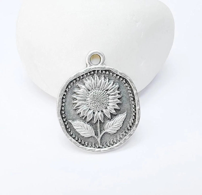 Silver-tone sunflower medallion charm with textured petals and leaf, round frame, hanging loop
