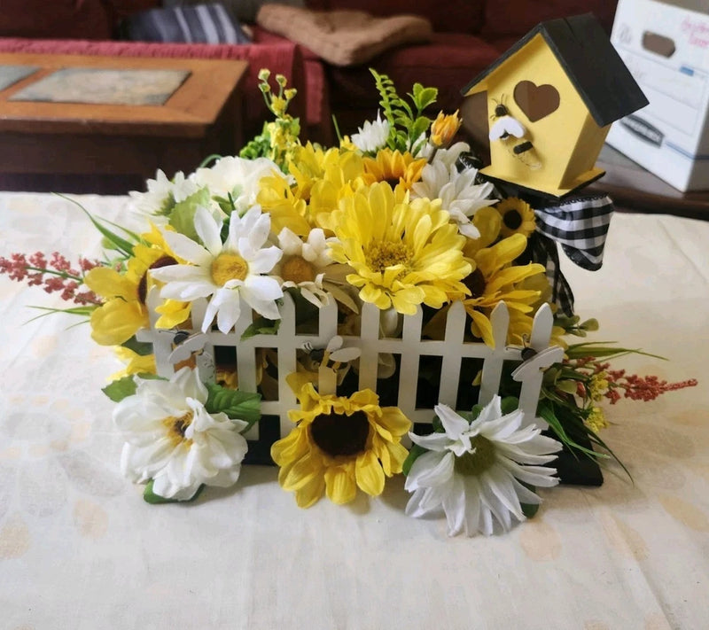 Artificial floral centerpiece with sunflowers, white flowers, and a wooden bee birdhouse