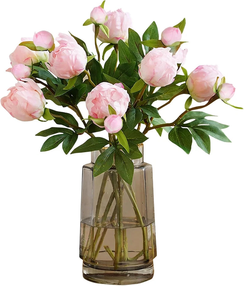 Pink artificial peony flowers with green leaves and stems in a clear glass vase