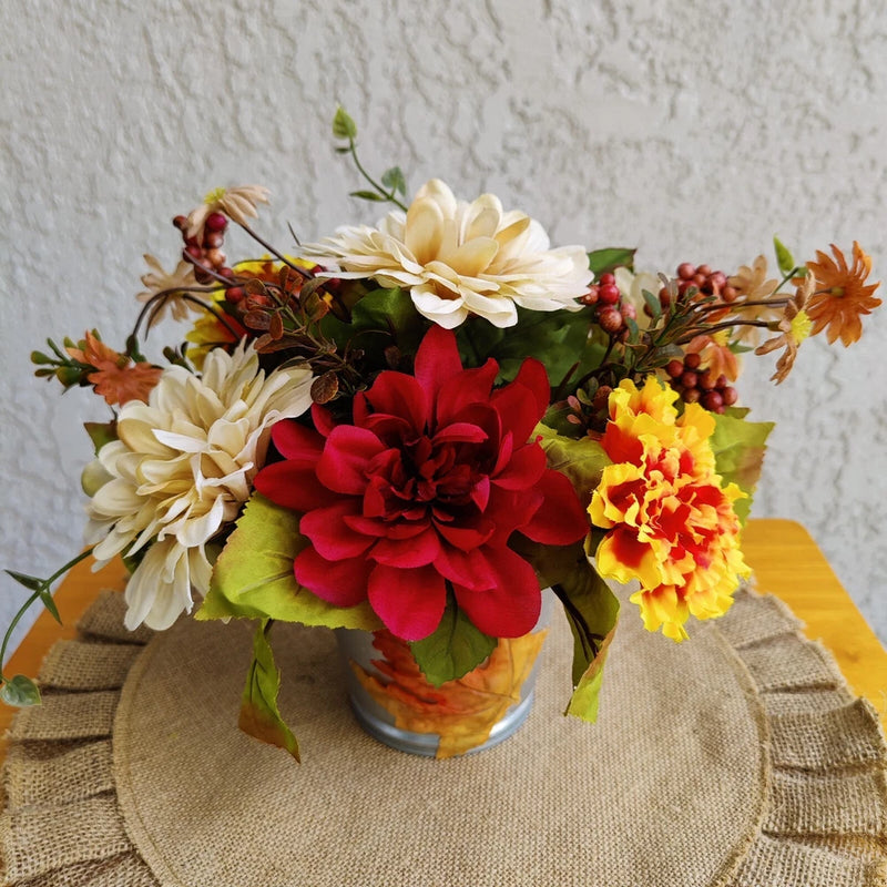 Autumn floral centerpiece in metal vase with cream, red, and yellow blooms on burlap runner