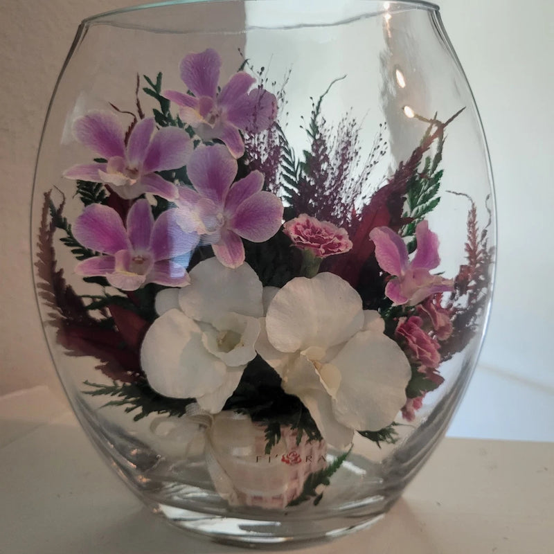 Glass-encased orchid arrangement with purple and white blooms and greenery