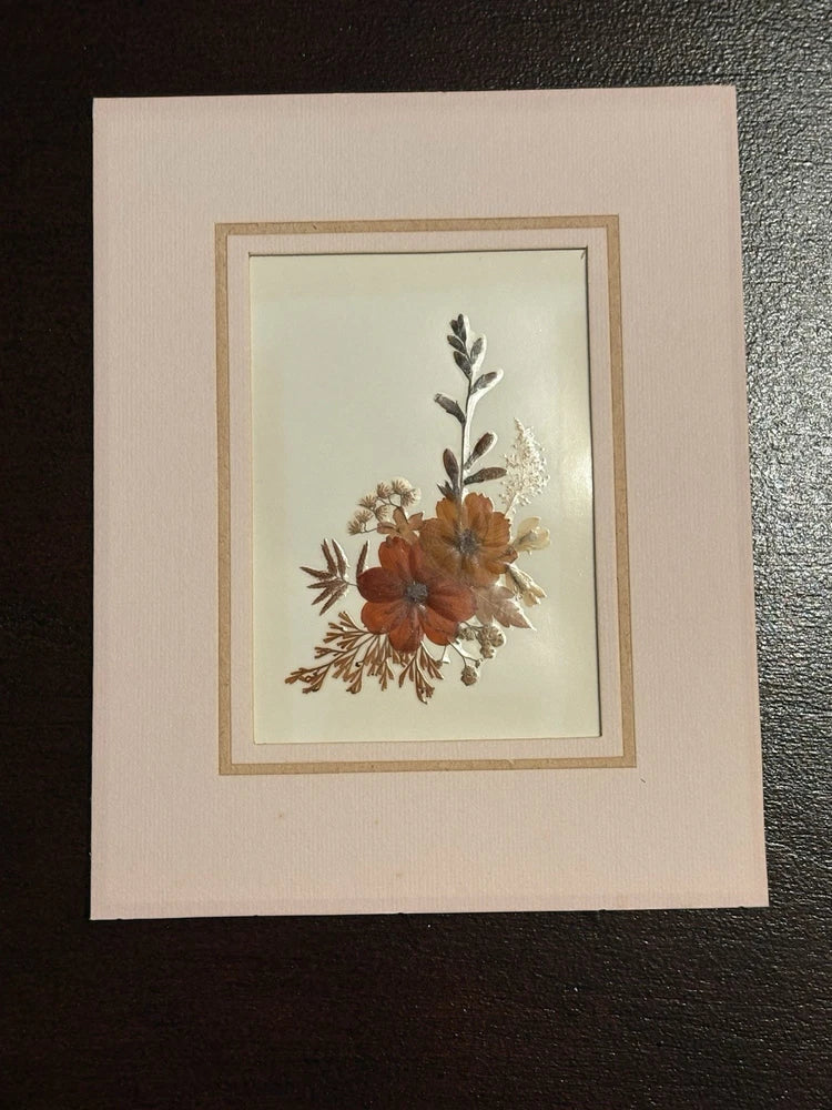Pressed flower art in gold-toned mat, featuring orange and cream dried blooms on light background