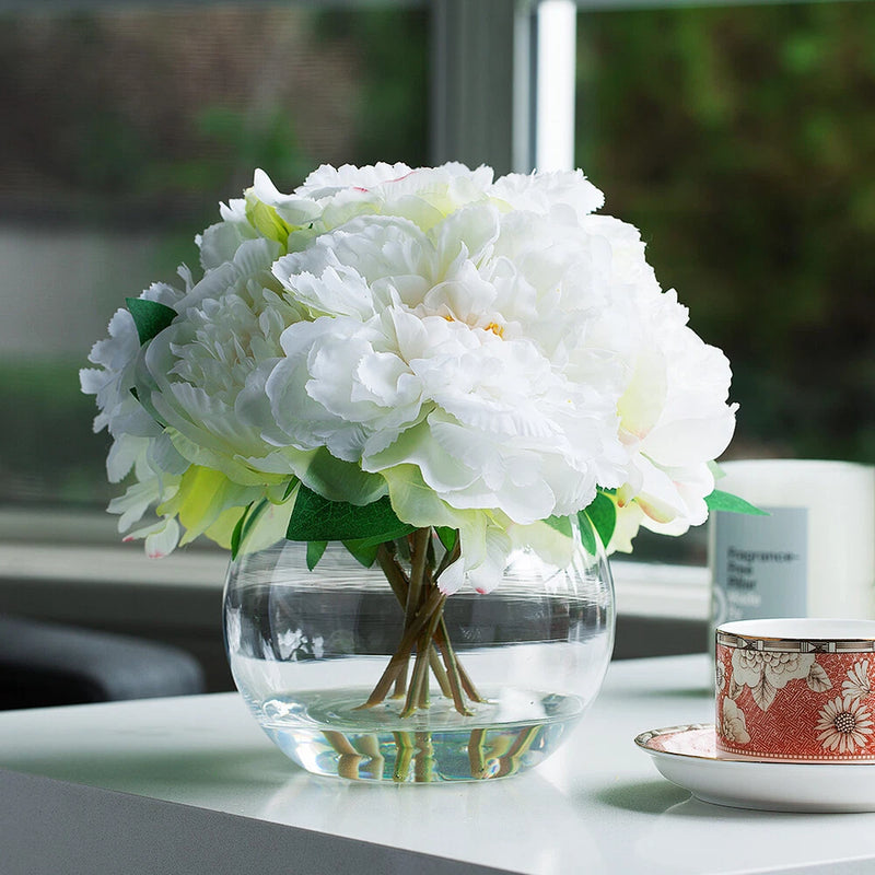 White artificial peony flower arrangement in round glass vase with green leaves