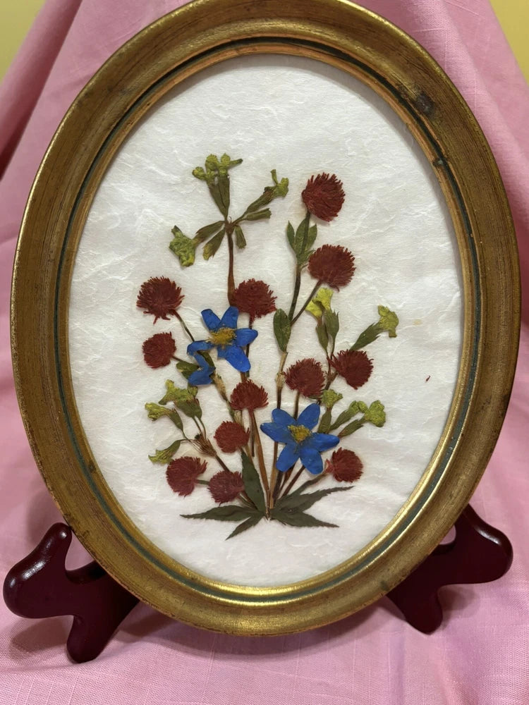 Oval gold frame with pressed flowers, red blooms and blue accents on white backing