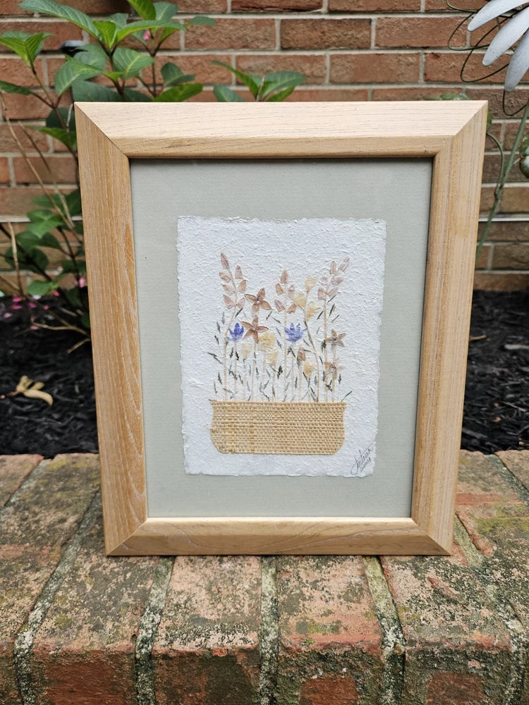 Framed pressed-flower wall art with blue blooms on white paper, in light wood frame