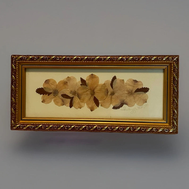 Framed pressed flower artwork with golden ornate frame, beige blossoms, and signature