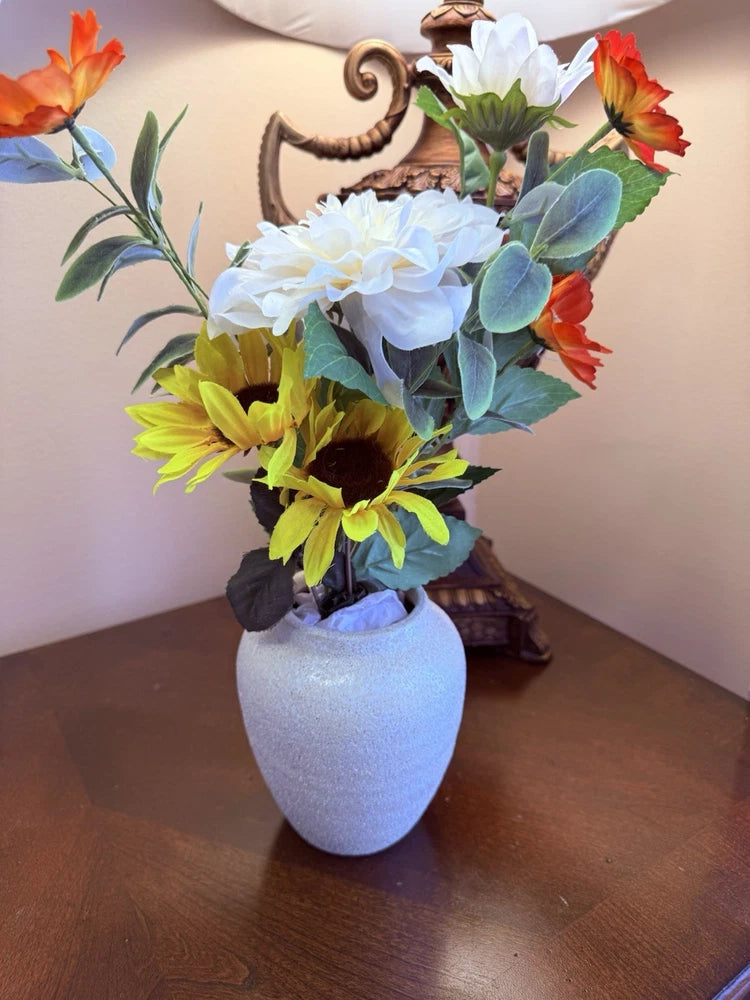 Artificial flower arrangement in a white ceramic vase with orange, yellow, and white blooms