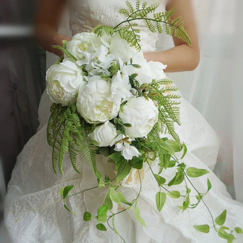 White artificial bride bouquet with peonies and leafy greenery, held by woman in white dress