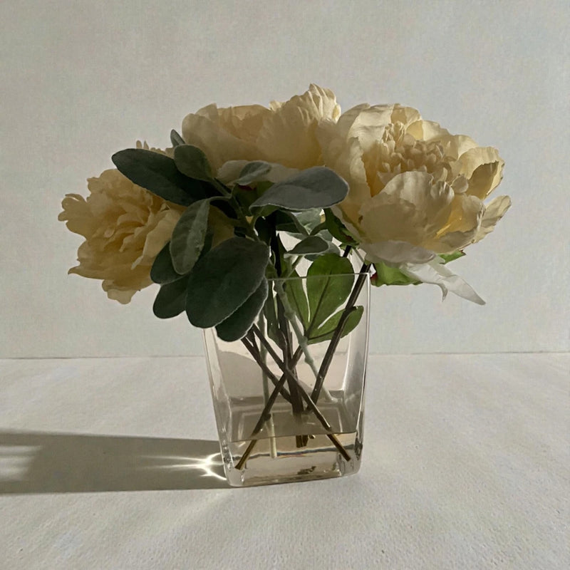 Artificial flower arrangement in clear cube glass vase with cream blooms and dark green leaves