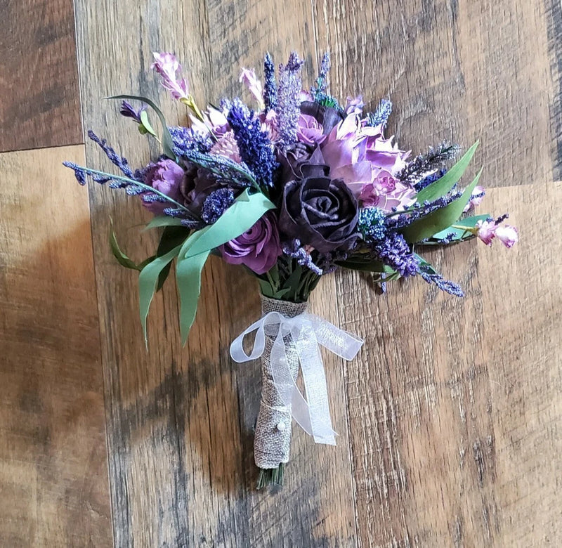 Lavender and purple bridal bouquet with rose blooms, teal leaves, and white lace ribbon handle