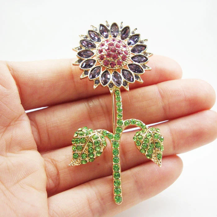 Gold-tone sunflower brooch with purple crystal petals, pink center, and green rhinestone leaves