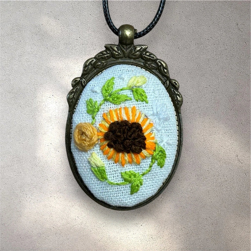 Embroidered sunflower pendant necklace with oval blue fabric, bronze frame, black cord