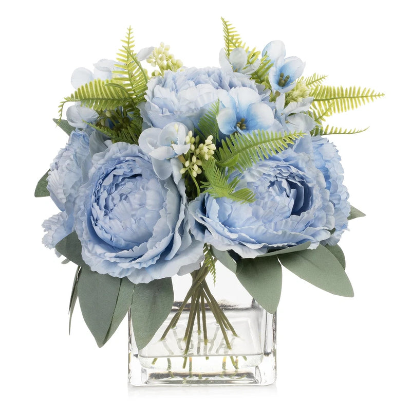Artificial peony flower arrangement with blue blooms and green leaves in clear glass vase