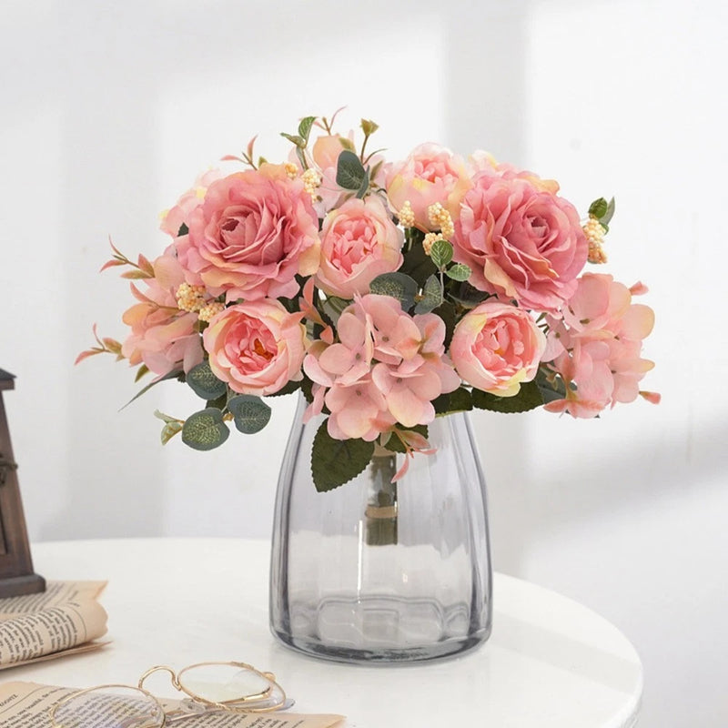Artificial pink peony flower arrangement in clear glass vase with green leaves