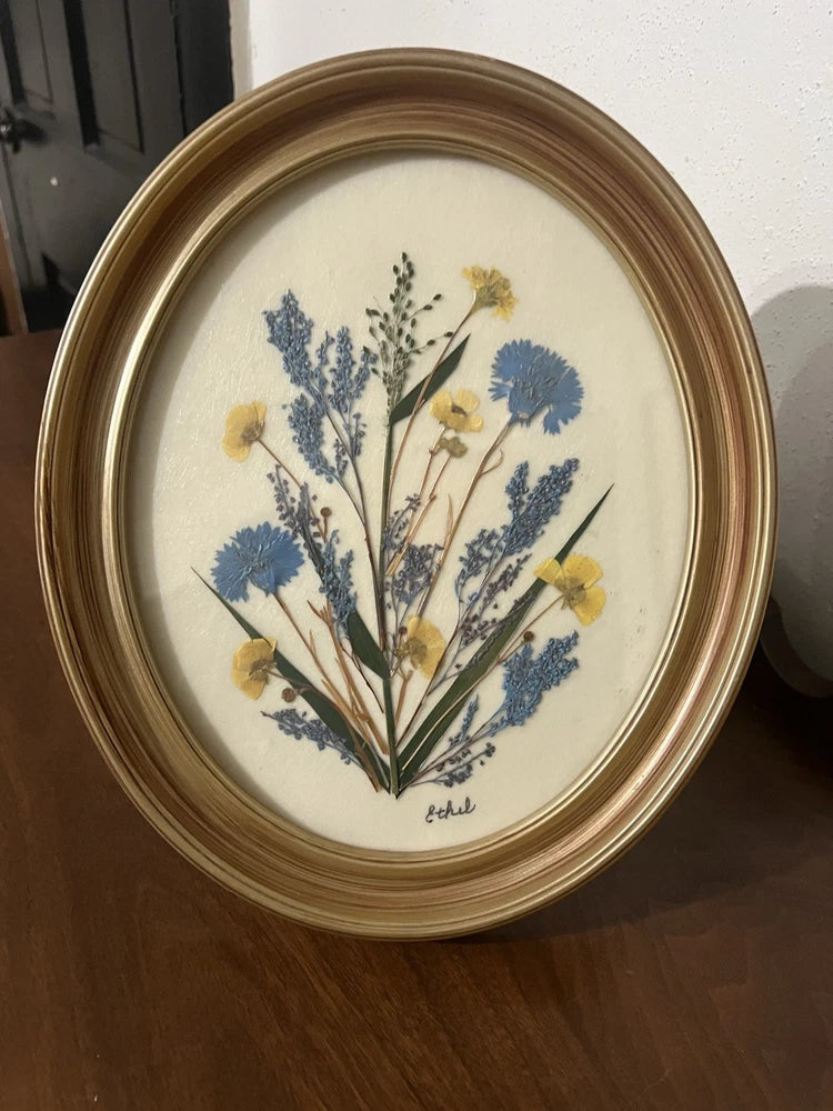 Oval wood frame dried flower art with blue and yellow pressed botanicals on light background